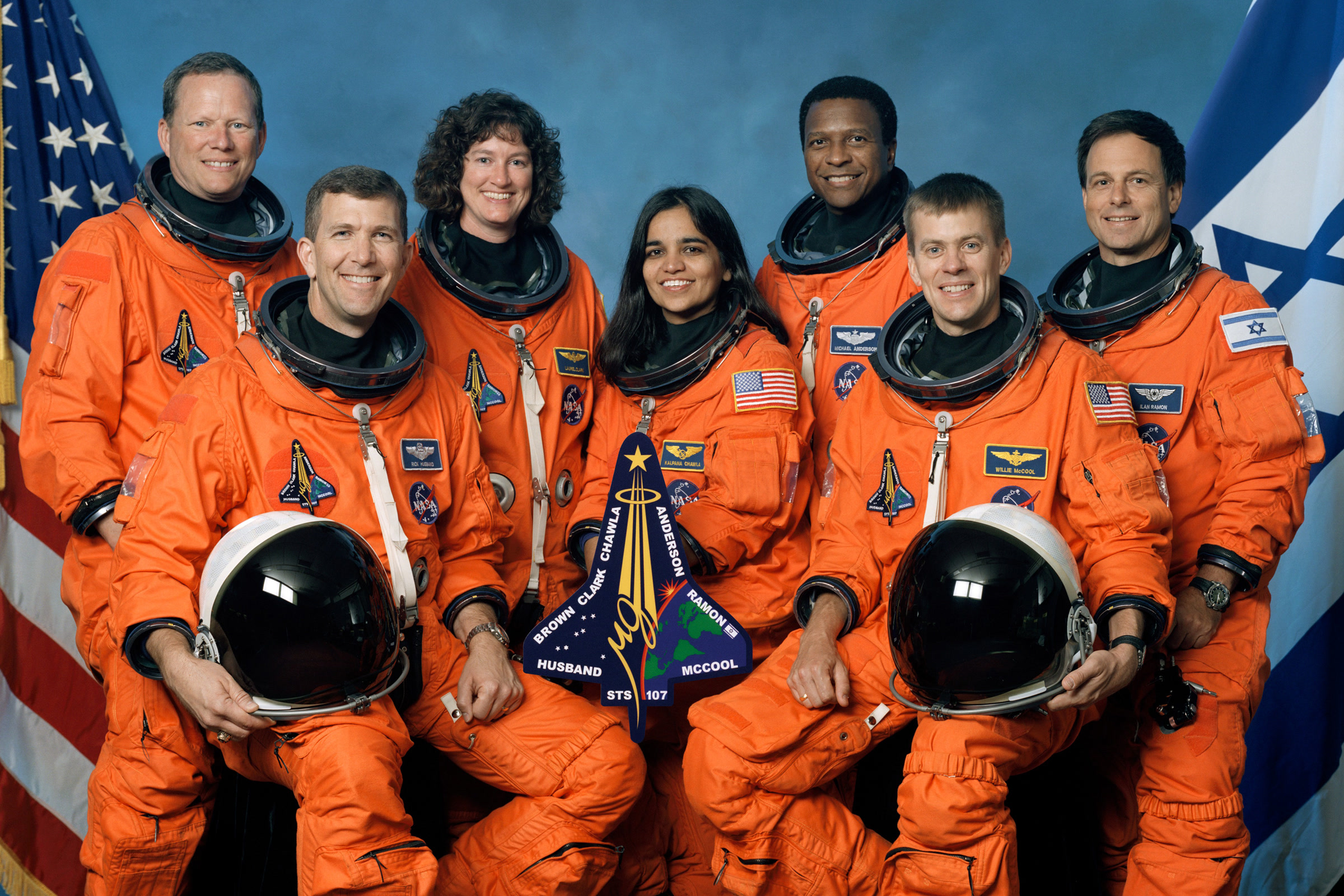 The seven STS-107 crew members in orange launch and entry suits pose around their mission patch, with Willie McCool seated at front right
