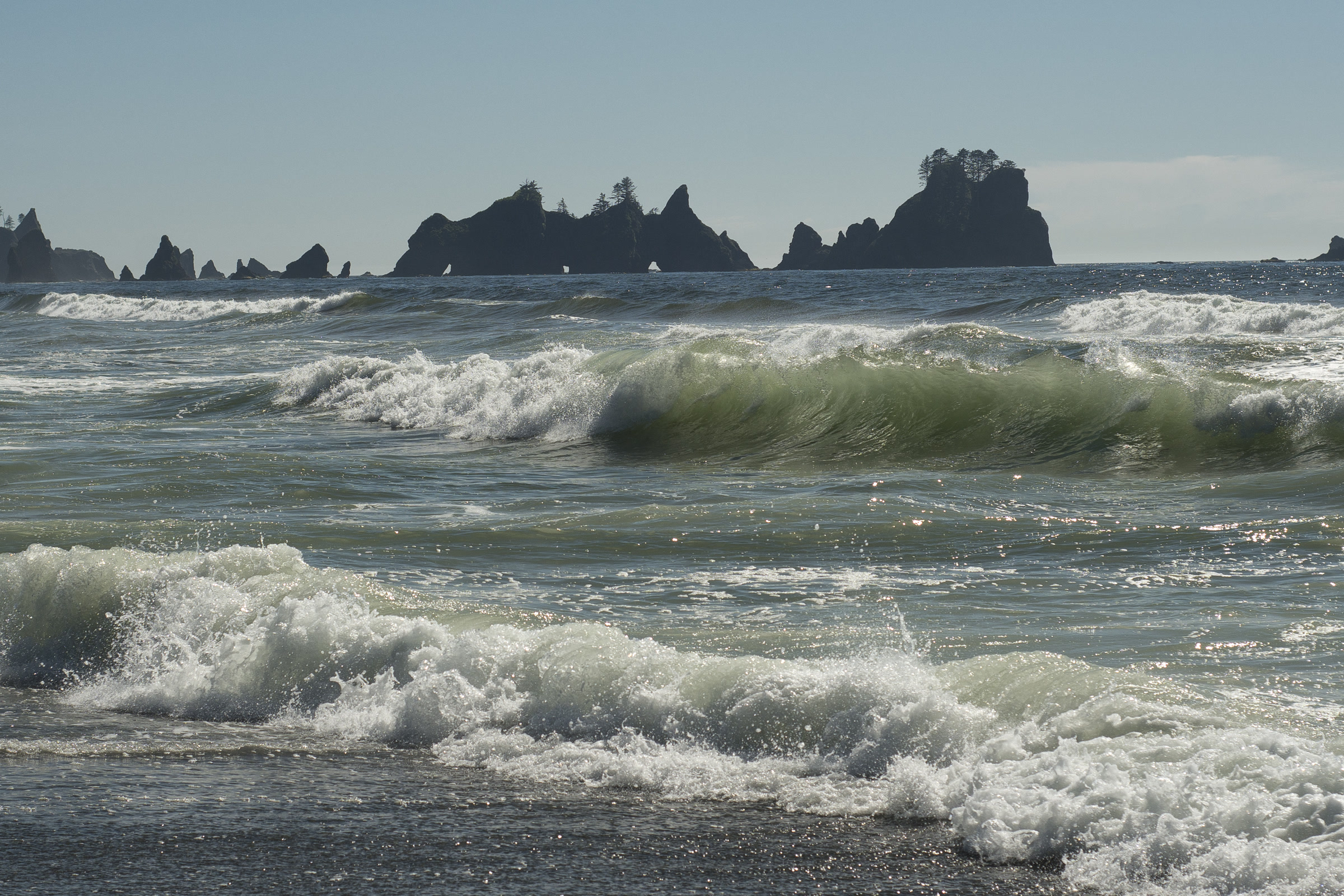 Green breaking waves rolling toward the shore of Shi Shi Beach, with the jagged sea stacks of Point of the Arches on the horizon