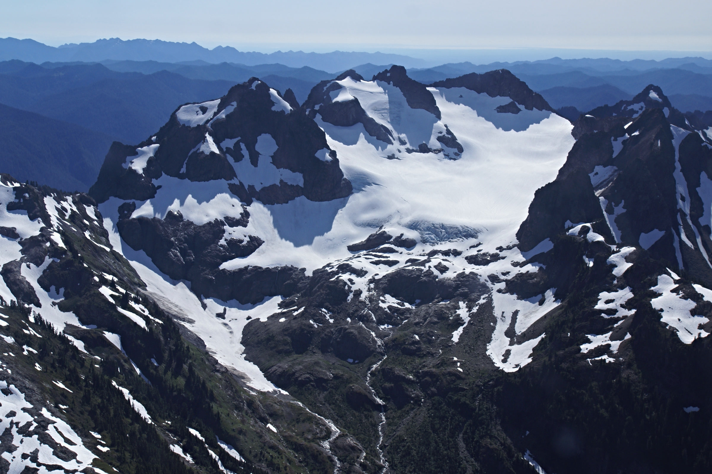 Aerial view of the rocky Valhallas peaks beside Mount Olympus, with a broad glacier and snowfields on their flanks