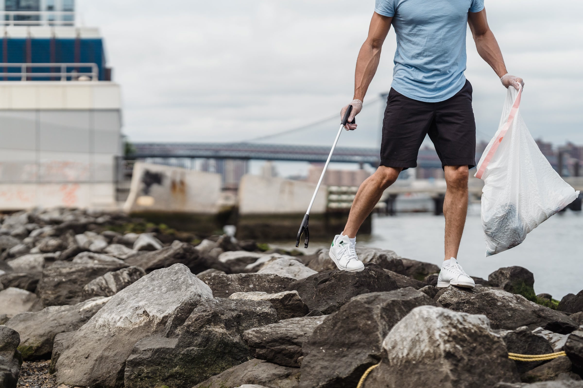 A volunteer in gloves, shown from the chest down, picking up litter with a grabber on riverside rocks and holding a trash bag