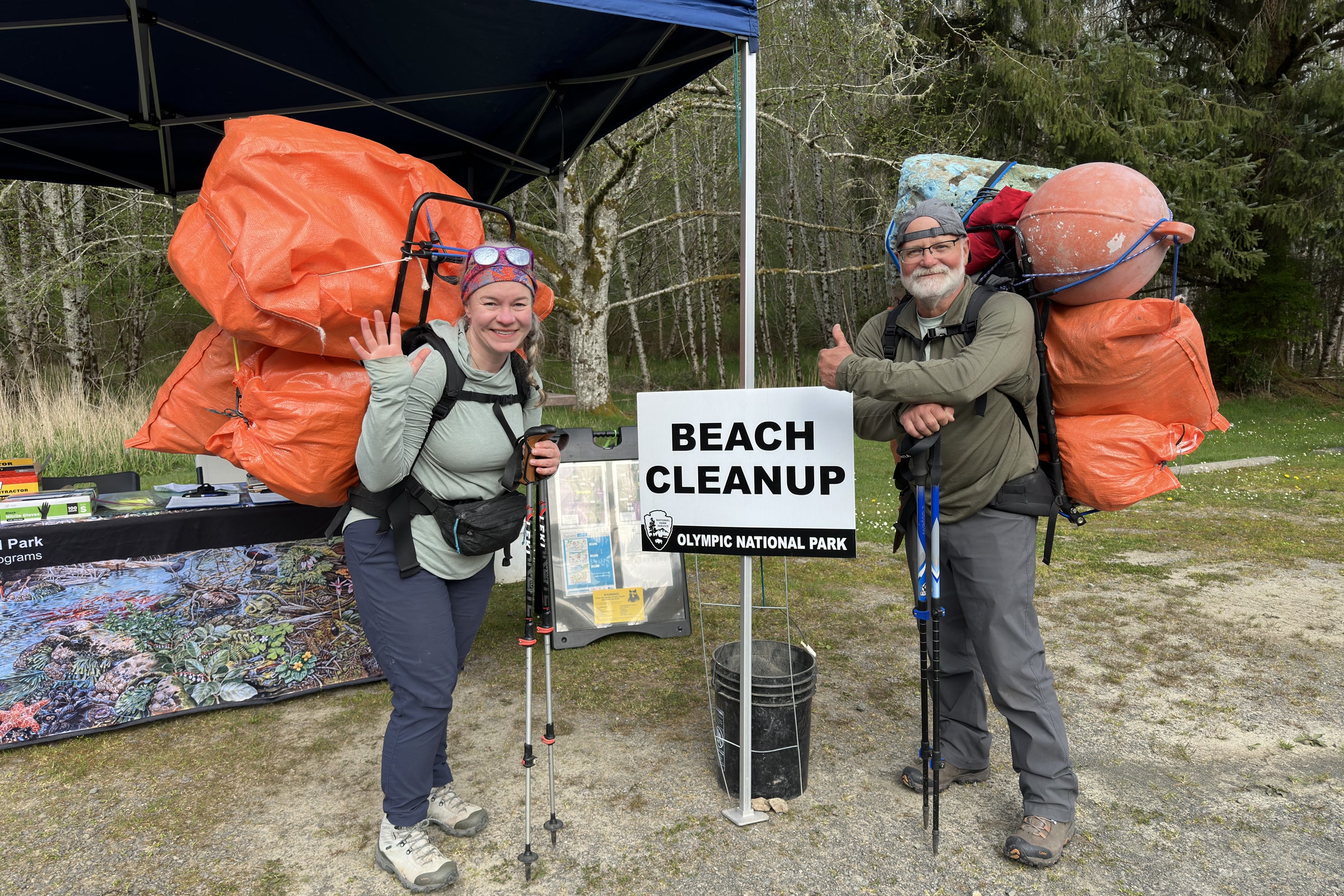 Two volunteers at an Olympic National Park beach cleanup station near Ozette, wearing backpacks stacked with bags of collected litter