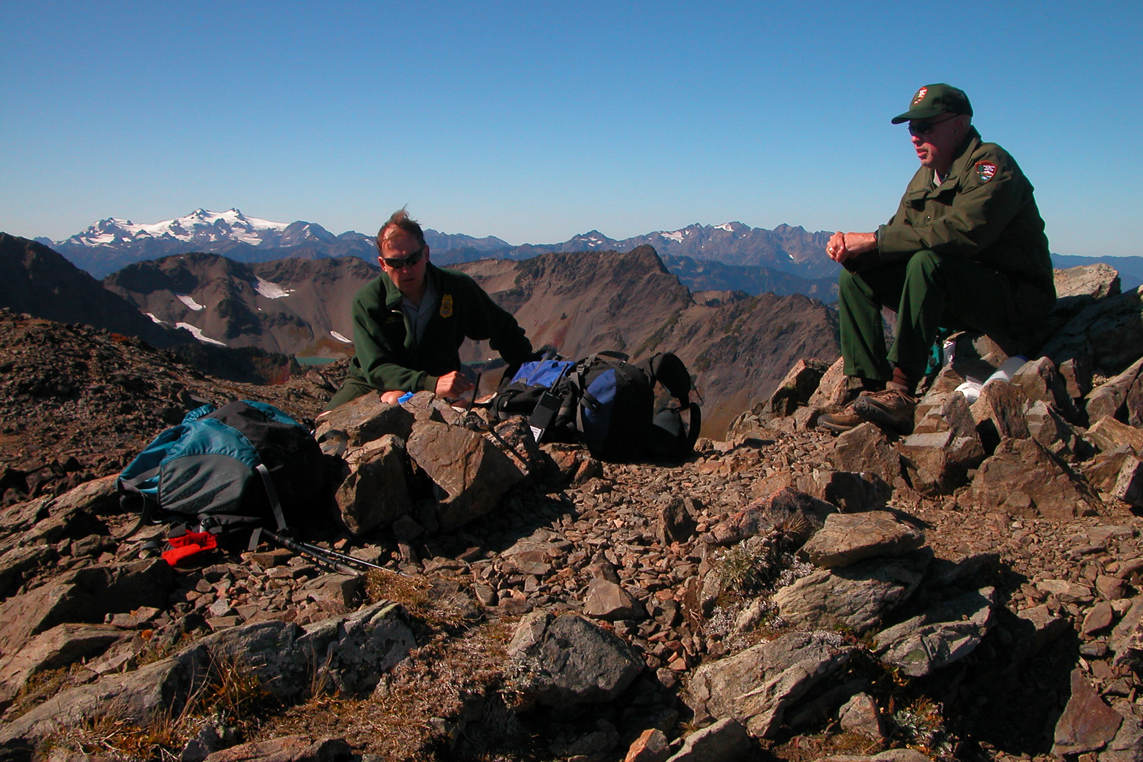 Two park rangers with backpacks resting on a rocky ridge with snowy Olympic peaks in the distance