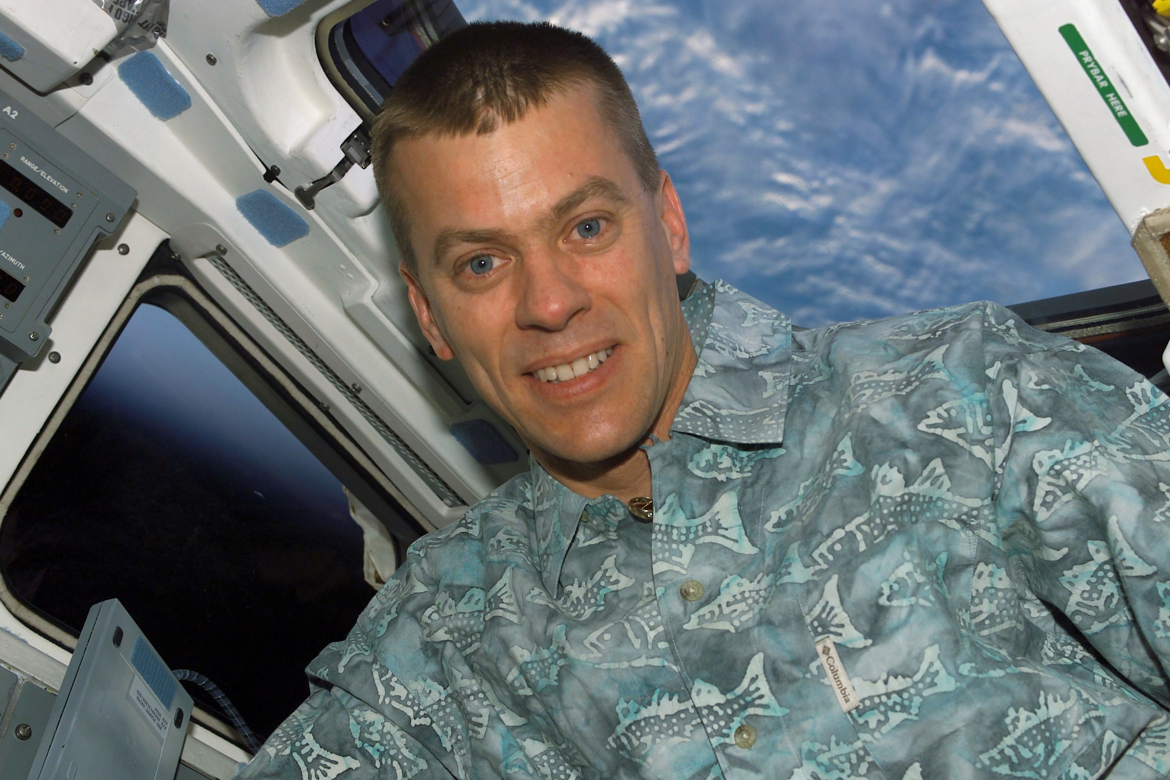 Astronaut Willie McCool smiles from the aft flight deck of Space Shuttle Columbia in orbit, with clouds over Earth visible through the window behind him