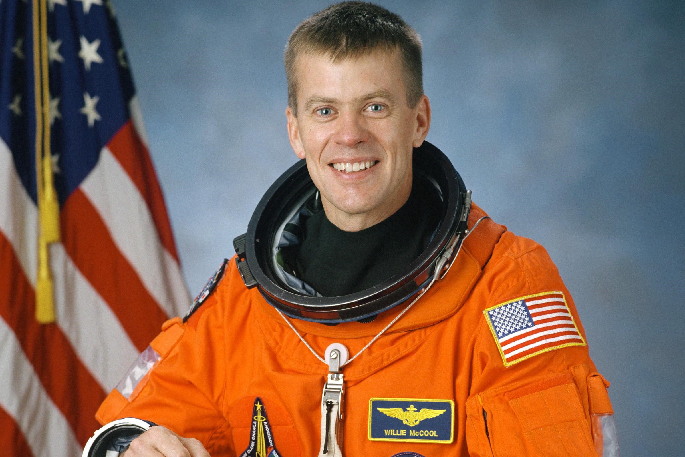 Official NASA portrait of astronaut Willie McCool in an orange launch and entry suit, smiling in front of an American flag