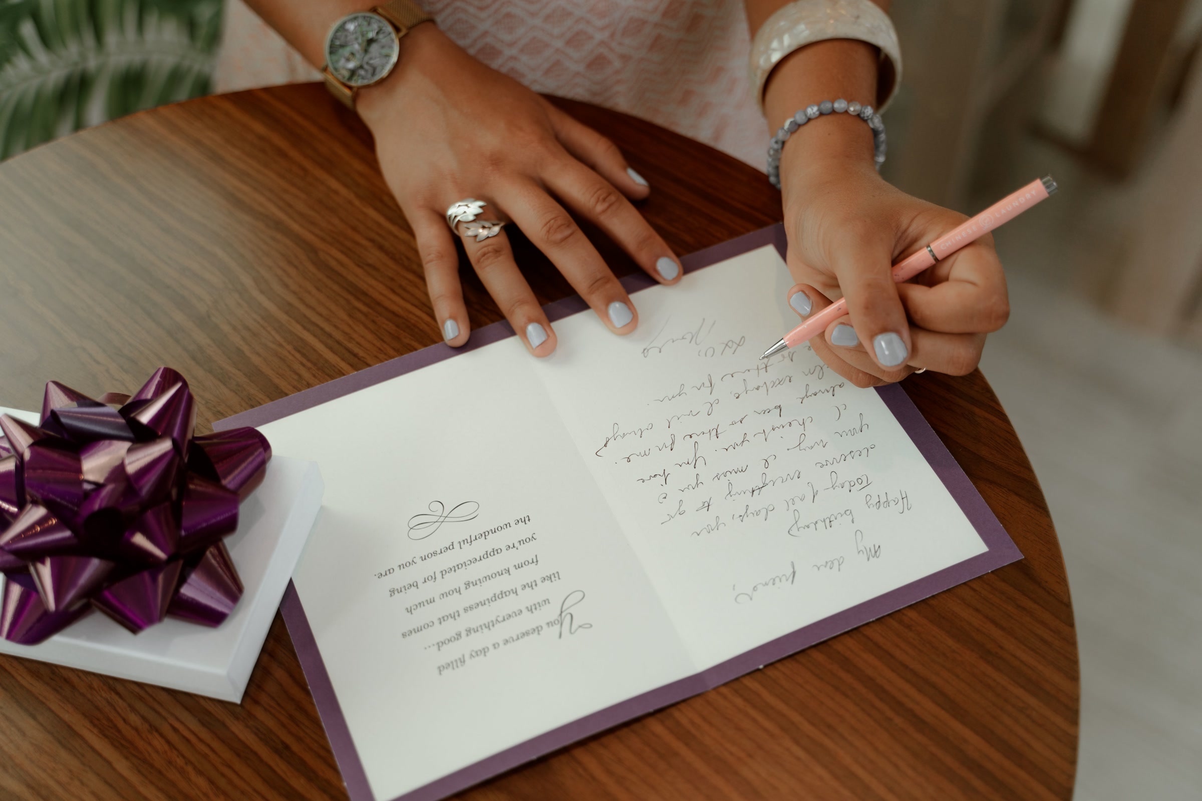 Hands writing a handwritten message inside a card on a wooden table, beside a small wrapped box with a purple bow