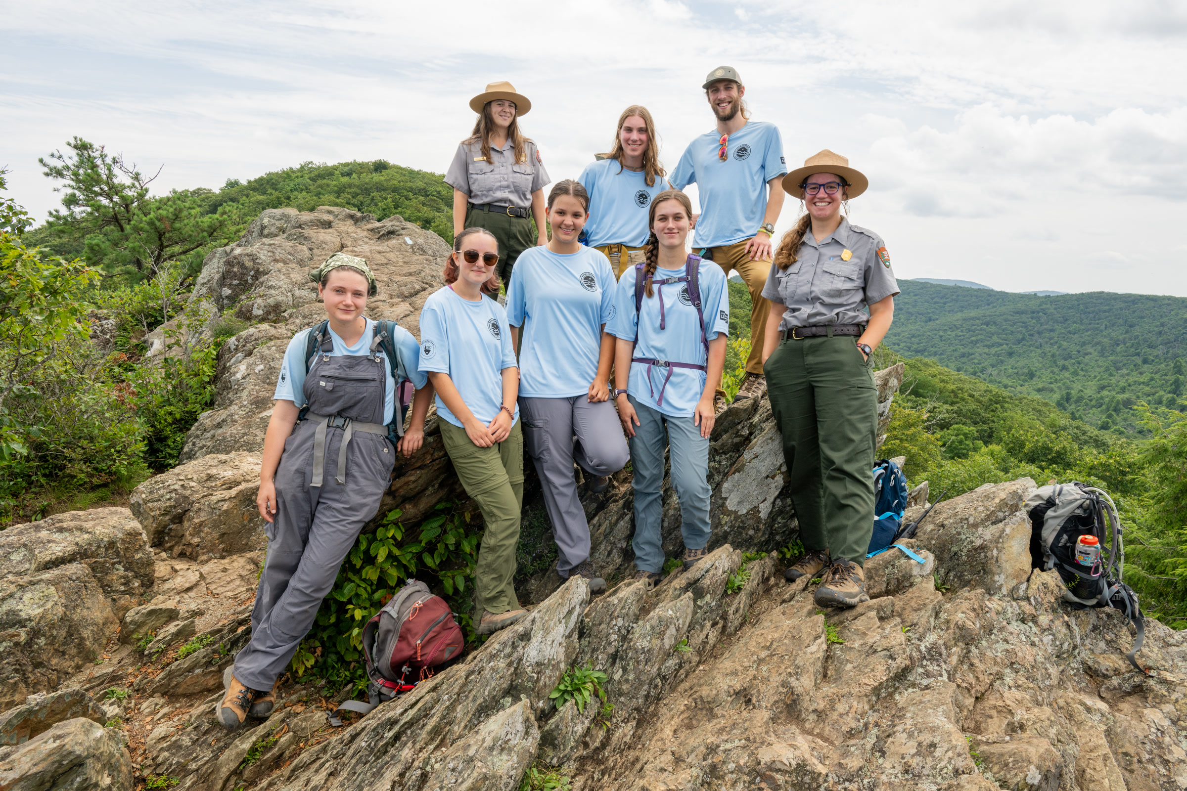 Seven young crew members in light blue shirts and a park ranger in a flat hat pose on a rocky outcrop above forested hills