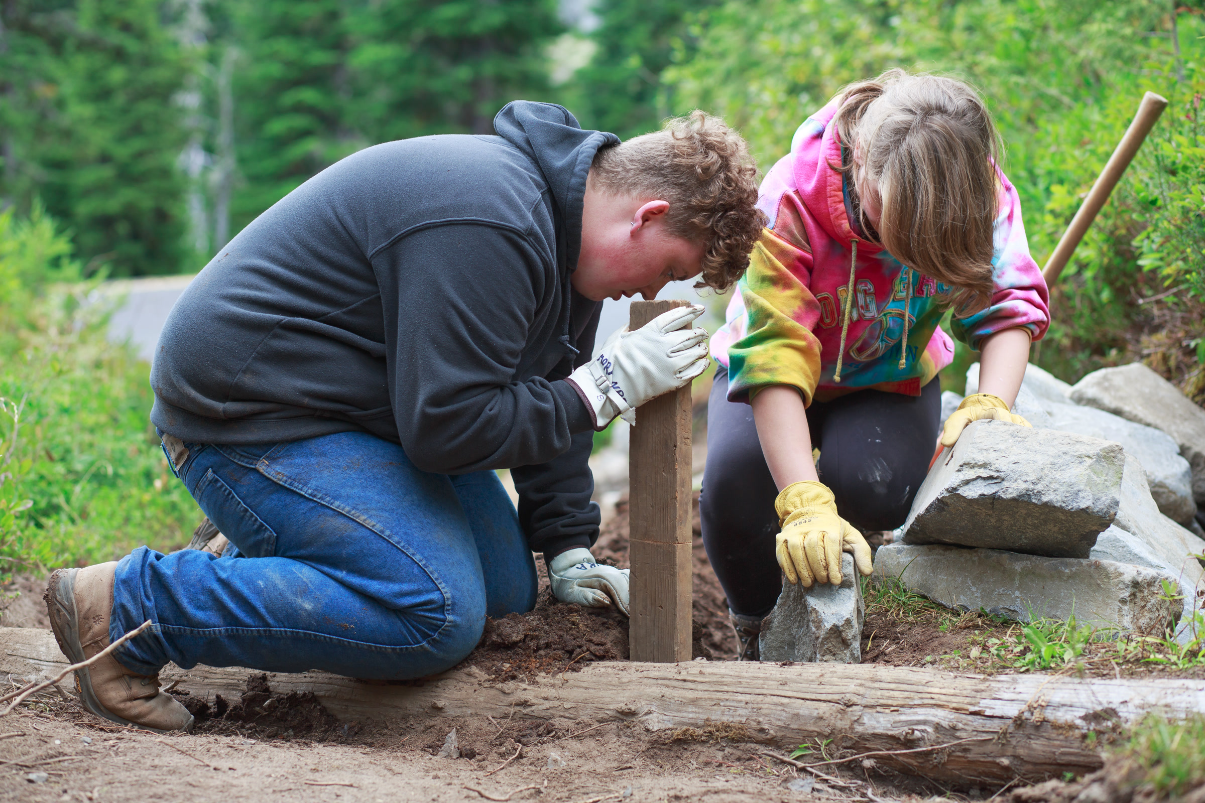 Two Youth Conservation Corps members in work gloves kneel to set rocks and a stake along the Lakes Trail at Mount Rainier
